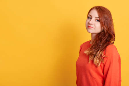 Portrait of young stunning model with red wavy hair, pretty woman in fashionable orange sweater, posing against yellow background. Copy space for advertisement or promotional text.の写真素材