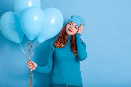 Calm young girl with closed eyes celebrating, putting hand on head on bright blue background with...
