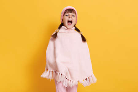 Young little girl dresses poncho and cap, standing against yellow wall and yelling happily, looks at...
