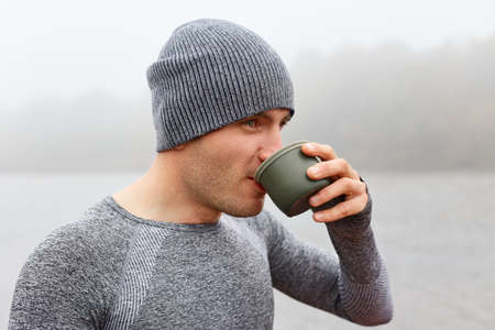 Young guy enjoying drinking coffee on river bank. Handsome man holding cup of tea outdoors. Morning...