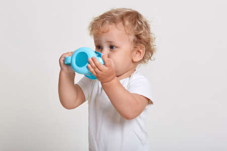 Little boy with blue plastic baby cup drinking water, thirsty child in t shirt posing isolated over...