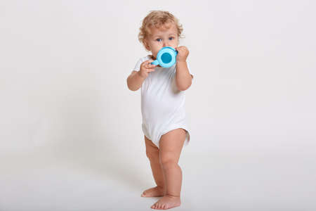 Blond curly haired baby drinks water from bottle, looks directly at camera, wearing bodysuit, full...