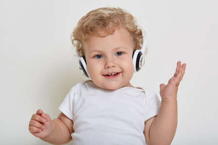 Beautiful little boy on white background with headphones, looking directly at camera with happy smile, showing hid teeth, wearing t shirt, blond curly haired kid with raising hand listens songs.の写真素材