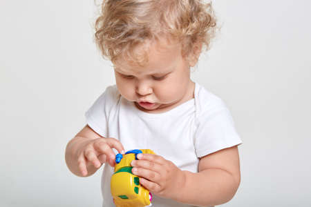 Little girl playing with yellow car, looking at toy car with great interest, wearing t shirt, posing...