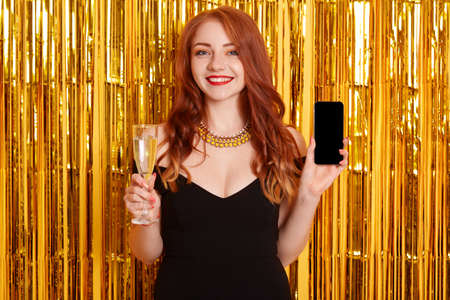 Smiling young woman in black dress and necklace, holding mobile phone with blank empty black screen and glass of champagne on bright background decorated of tinsel, birthday, holiday, party.の写真素材