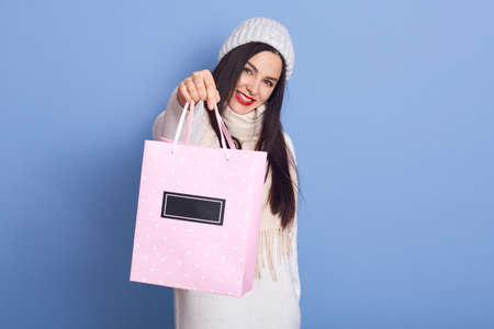 Winsome young brunette woman girl in warm white sweater posing isolated on blue background, holding...