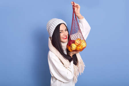 Cheerful laughing woman standing with fruits in net bag isolated over blue studio background, female...