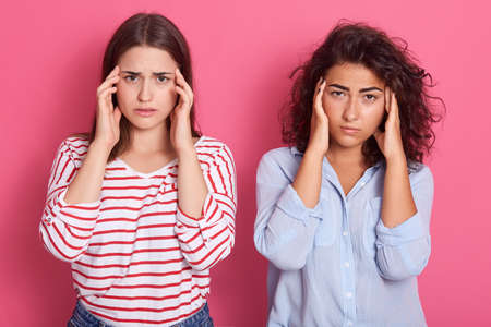 Tired dark haired women friends have headache, look in displeasure from forehead, keep hands on...