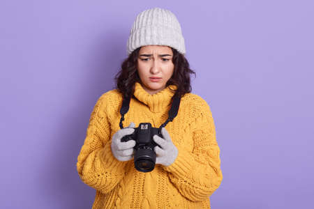 Woman with photographic camera standing against lilac background, has scared facial expression, sees...
