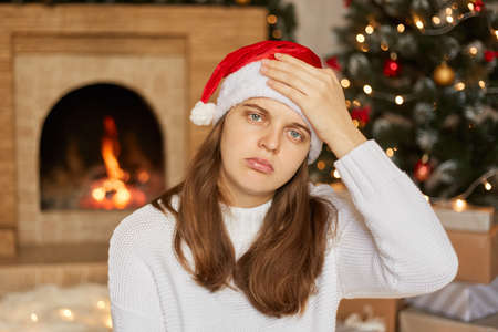 Stressed christmas woman having headache, keeping palm on her forehead, looks sick and unhappy, looking directly at camera, suffering from cephalagia, fireplace and x-mas tee on background.の写真素材