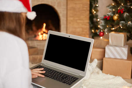Photo of charming lady sitting on floor and holding laptop with blank screen, wear red santa claus...