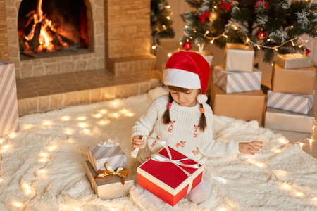 Cute little girl opening Christmas gift at home while sitting on soft carpet on floor, looking at box, pulls ribbon to open present, dresses white pullover and red santa hat.の写真素材