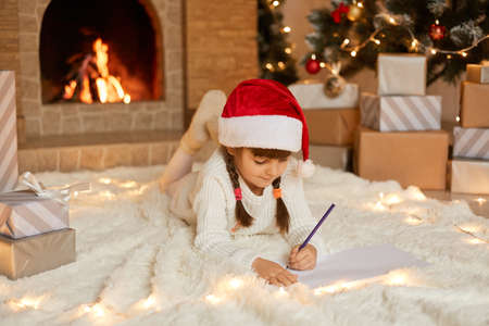 Little cute female child lying on floor on carpet and writing letter to Santa Claus, looking at empty paper and think, posing against present boxes, xmas tree and fire places.の写真素材