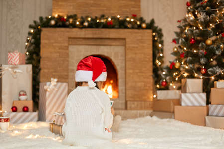 Child looking at smart phone or tablet screen while sitting near fireplace on floor, posing backwards, wearing white sweater and santa hat, posing in living room with new year decoration.の写真素材
