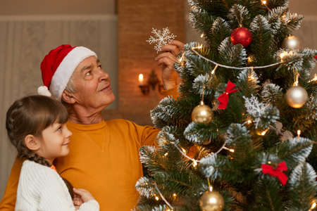 Side view of grandfather with his little granddaughter decorating New Year tree, man holds christmas...