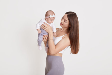 Cheerful beautiful young woman in bra and gray leggins, holding baby girl in her hands and looking at her, brown haired female posing with child against white wall.の写真素材