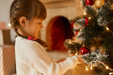 Kid decorating Christmas tree, charming girl with pigtails wearing white sweater, looking at fur tree with garland, posing in festive room, child waits New year tree.の写真素材