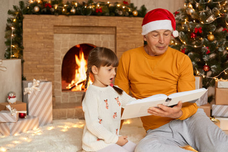 Grandfather and granddaughter reading book near fireplace and xmas tree, grandpa and grandchild with astonished facial expression, family spending new year together, reading Xmas stories.の写真素材