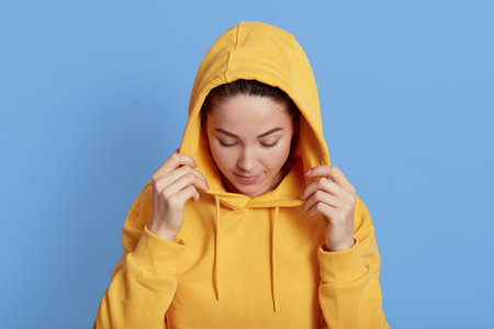 Beautiful girl wearing yellow hood, looking down, having serious expression, standing isolated over blue background, attractive girl keeping hand on hood.の写真素材