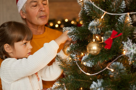 Happy family decorating xmas tree, grandfather wearing orange sweater and little girl in white jumper posing in living room, beautiful xmas tree with bubbles and bows.の写真素材