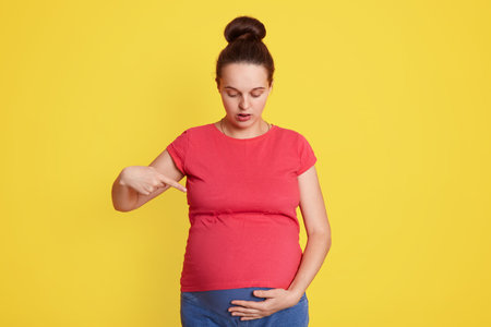 Attractive young pregnant female with astonished facial expression, looking at her belly and looks at it, pointing with index finger on her tummy, wearing red t shirt, stands isolated over yellow wallの写真素材