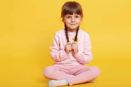 Full body photo of coy kid sitting with crossed legs, wearing casual outfit isolated over yellow background, keeping hands on her pigtails, looks at camera with timid expression.の写真素材