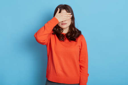 Portrait of young pretty Caucasian scared woman covering eyes with hands while standing against blue background, brunette exhausted girl close eyes with palm ignoring something.の写真素材
