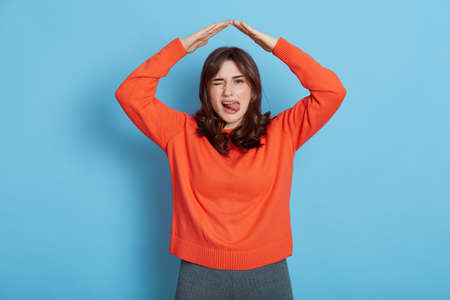 Portrait of happy girl in orange shirt feeling safe, confident under house, making roof gesture, smiling and showing tongue, looking at camera and blinking one eye.の写真素材
