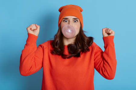 Excited dark haired female blows bubble from chewing gum, has amazed expression, dresses casual clothes, model posing isolated over blue background, cant believe in her success, clenches fists.の写真素材