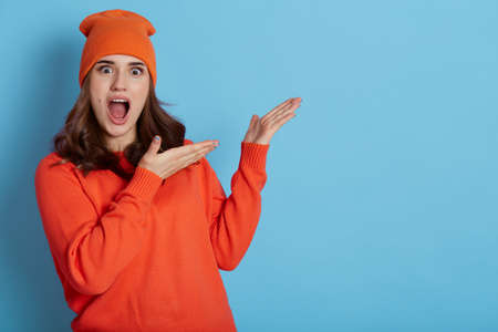 Horizontal shot of joyful Caucasian female with overjoyed expression, showing something amazing aside against blue background, dressed in casual clothing, copy space for advertisement.の写真素材