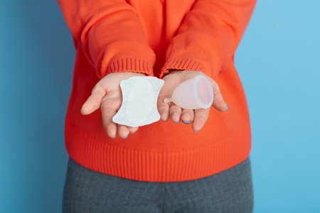 Unknown female holding hygiene pad and menstrual cup in both hands, not knowing what to choose, faceless girl in casual clothing holds personal hygiene products isolated over blue background.の写真素材