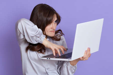 Angry executive elegant job woman typing on laptop, mad female working online, has lots work, looking at device screen with annoyed expression, wearing elegant blouse against lilac wall.の写真素材