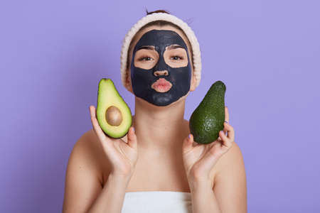 Beautiful young European woman with black facial mask and fresh avocado in hands isolated on lilac background, making kissing gesture with lips, looks at camera, skin care procedures for cleansing.の写真素材