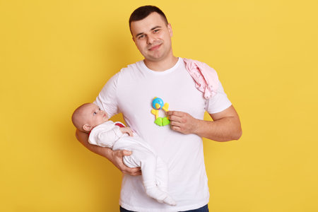 Smiling man wearing casual attire stands against yellow wall with his newborn baby in handsの写真素材