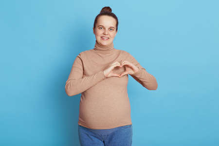 Smiling happy future mother standing against blue wall, looking directly at camera, has hair bun, wearing casual beige sweater and jeans, making heart gesture in front of chest, expressing love.の写真素材