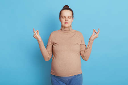 Peaceful pregnant woman meditating isolated over blue background, keeping eyes closed and raising...