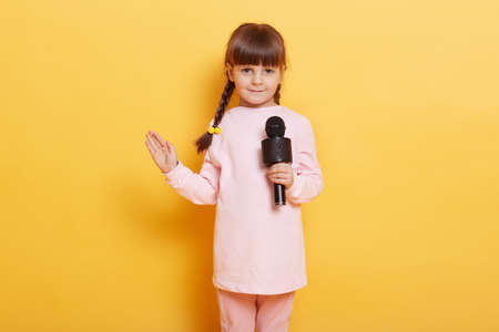 Girl singing with microphone and waving palm to camera, smiles, looking cute and charming, looks at camera, wearing casual clothing, kid with pigtails arranging concert for somebody.の写真素材