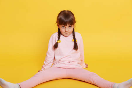 Beautiful small European girl sitting on floor and looking at camera from under forehead, female child wearing pink shirt and sweater, kid with pigtails isolated over yellow background.の写真素材