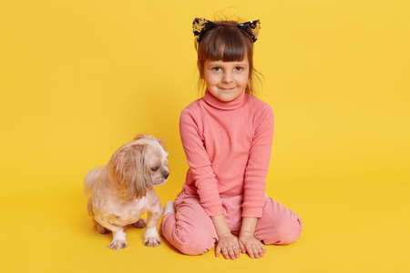 Girl with puppy isolated over yellow background, child looking directly at camera with smile, kid wearing rose sweater, pants and hair band with cat's ears, small kid with Pekingese.の写真素材