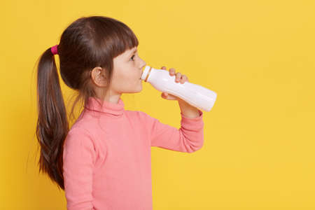 Child drinks milk while posing isolated over yellow background, profile of little dark haired female kid drinking yogurt from bottle, small girl wearing pink clothing, has ponytail, copy space.の写真素材