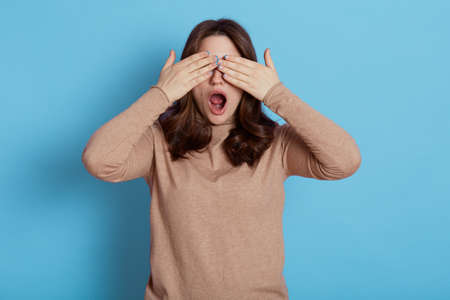 Shocked worried scared dark haired young woman years casual beige turtleneck, posing isolated on blue background, covering eyes with hands, hiding, keeps mouth widely opened.の写真素材