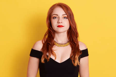 Serious sad ginger female, looking directly ar camera, woman with red lips wearing black elegant dress and golden necklace, posing isolated over yellow background.の写真素材
