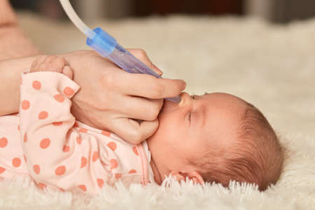 Faceless mother using nasal aspiration sucks the snot from baby, child lying on bed on fluffy blanket with closed eyes, unknown person taking care of infant.の写真素材
