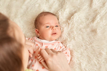 New born charming baby lying in bed with mother, looking away, studying world around, wearing...