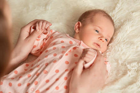 Close up portrait of newborn baby looking away, lying in bed on fluffy blanket, unknown person holding infants hands, faceless mother with her new born kid.の写真素材