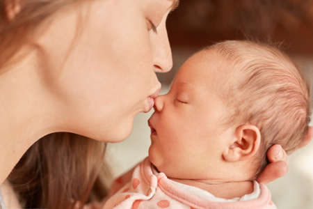 Mom with newborn baby posing indoor, mother kissing her sleepy infant, female closed eyes and pout...