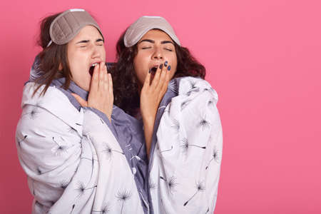 Two friends standing together over pink background, yawning and covering mouth with hands, restless and sleepiness, dark haired ladies with closed eyes, woman wrapped in blanket.の写真素材
