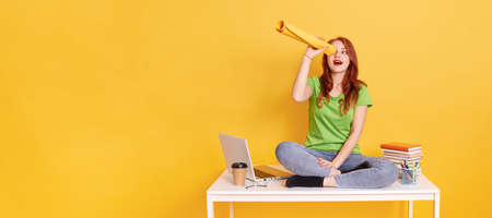 Playful young woman or student sitting on white table with crossed legs and twist paper, watching up though it like a binocular, keeps mouth opened, sees something curious, copy space for ad.の写真素材