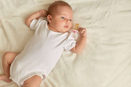 Tiny baby holding nipple on finger, looking away while lying on bred on white blanket, kid wearing...