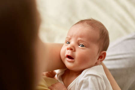 Woman posing backwards with newborn baby in hands, tiny girl or boy looks at mommy with curious expression and a bit opened mouth, wearing white clothing.の写真素材